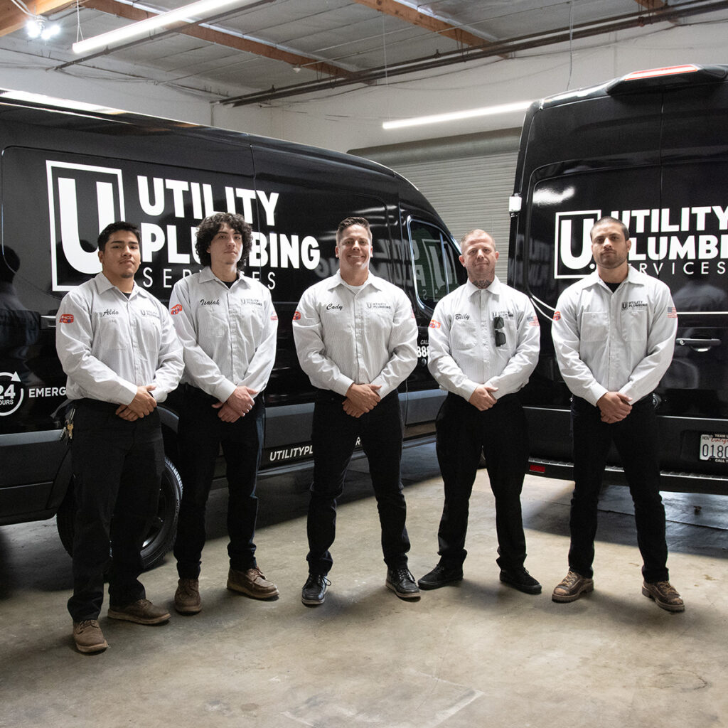 Five Utility Plumbing Services team members standing together in a warehouse in front of two branded service vans