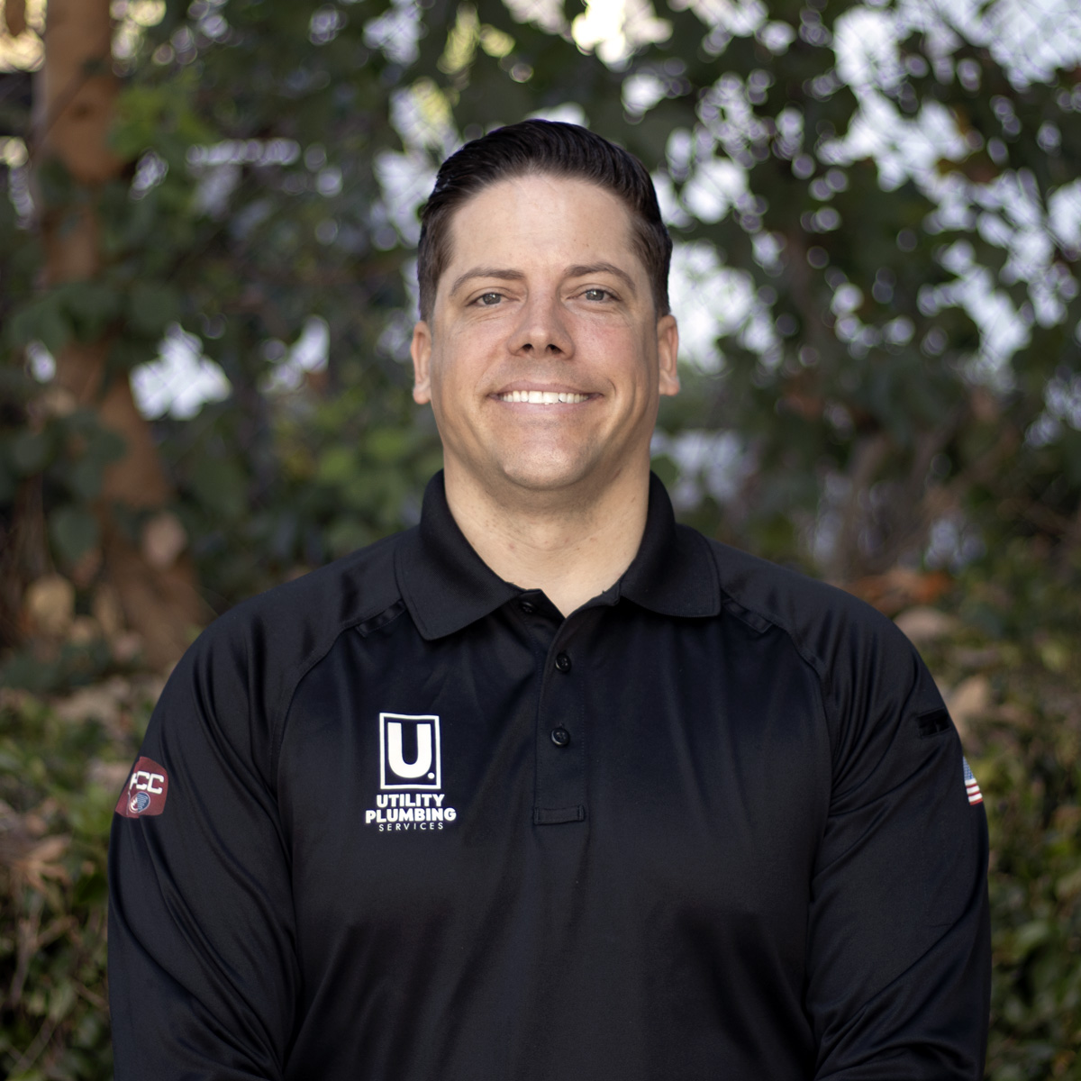 Smiling Utility Plumbing Services team member in a black logo polo poses for an outdoor headshot in front of green trees.
