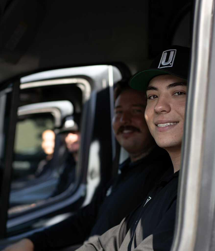 Smiling Utility Plumbing Services plumber in a black U cap sits in a work van beside a coworker in Rancho Cucamonga.